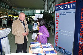 Ein Mann und eine Frau führen ein Gespräch in der Abflughalle vor einem Polizei-Plakat