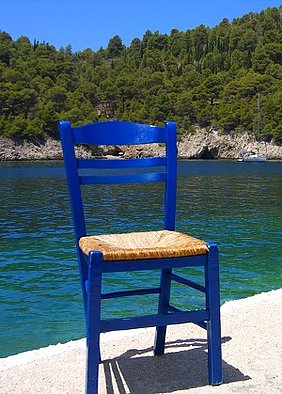 Blauer Stuhl mit heller Sitzfläche, im Hintergrund eine Bucht mit Meer und Bäumen