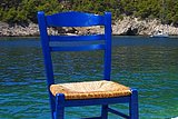 Blauer Stuhl mit heller Sitzfläche, im Hintergrund eine Bucht mit Meer und Bäumen